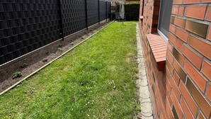 Un jardin avec un mur en briques, une clôture noire et une zone herbeuse.