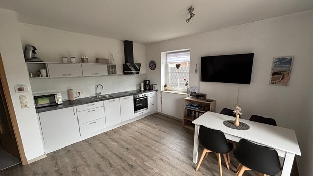 Une cuisine moderne avec des meubles blancs, un coin repas comprenant une table blanche et des chaises noires, et un téléviseur à écran plat fixé au mur.