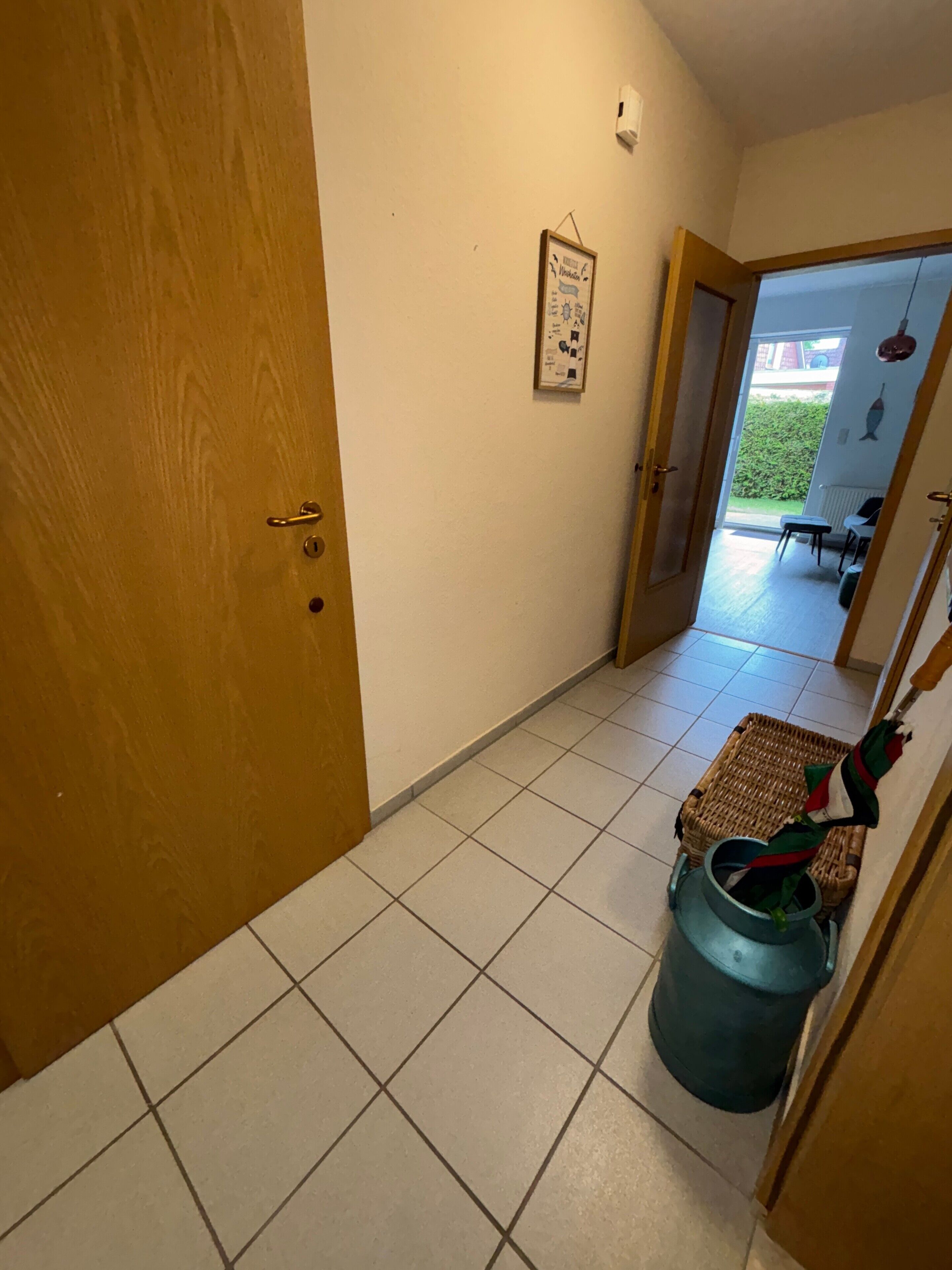 Un couloir avec une porte en bois, un tableau encadré au mur, et une chaise en osier, avec une poubelle et un parapluie à proximité.