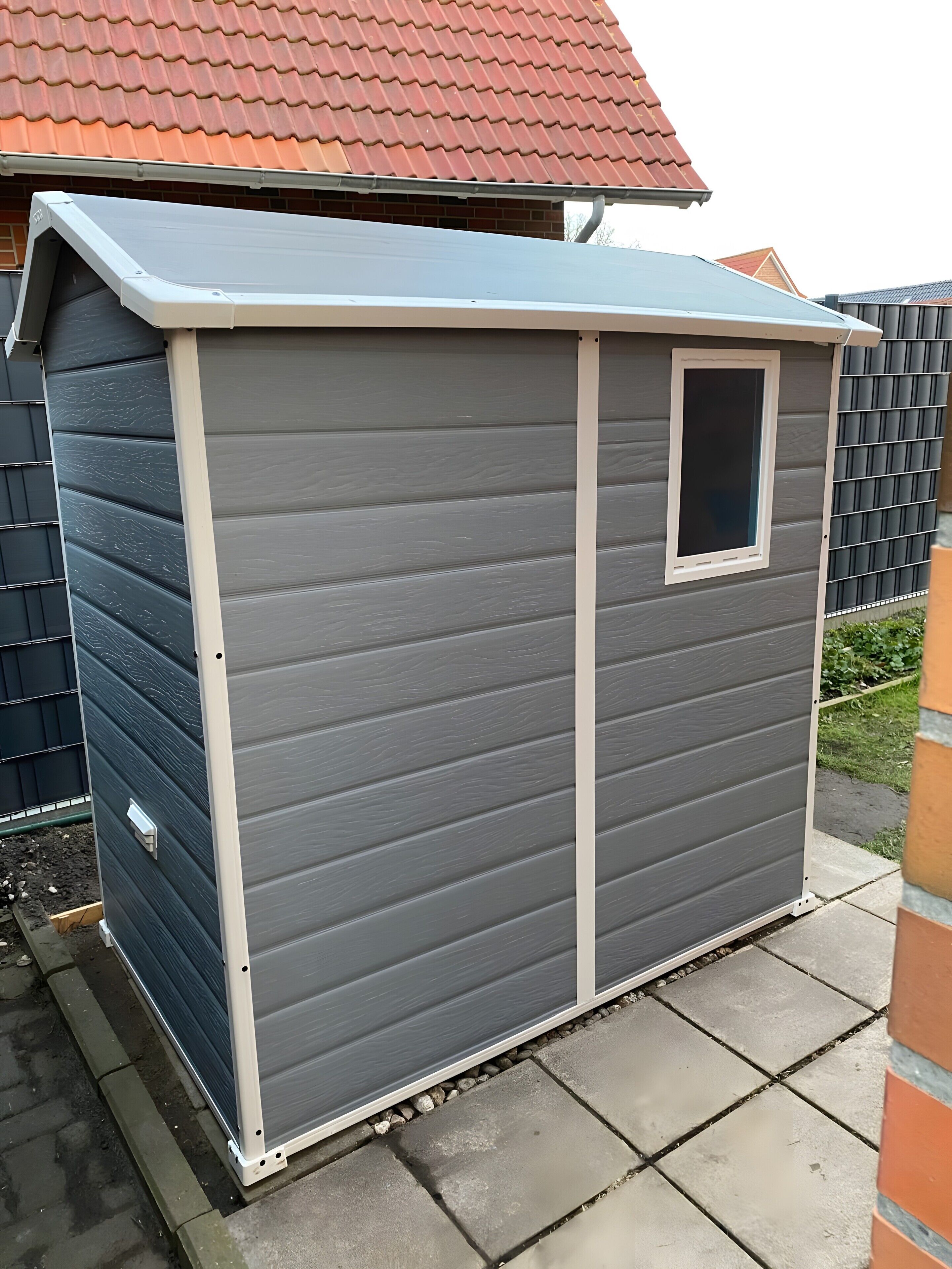 Un petit abri de jardin d’un seul étage, à l’extérieur gris et aux bordures blanches. Il possède une petite fenêtre à l’avant et est situé sur une terrasse dallée.