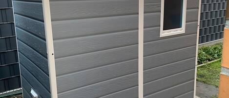 Un petit abri de jardin d’un seul étage, à l’extérieur gris et aux bordures blanches. Il possède une petite fenêtre à l’avant et est situé sur une terrasse dallée.
