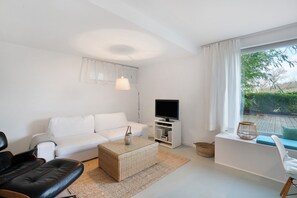 A modern living room with a white sofa, a black leather chair, a woven coffee table, a flat-screen TV on a white stand, and a large window with a view of greenery. - Apartment 'Modernes  Ammersee' with Private Terrace, Private Garden and Wi-Fi (Windach)