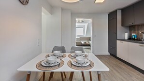 Ein moderner Essbereich mit einem weißen Tisch, gedeckt für vier Personen, einer Wanduhr und einer Küche im Hintergrund.