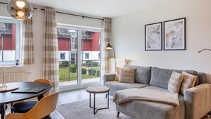 Un salon moderne avec un canapé, une petite table ronde et une table à manger avec des chaises. Il y a une grande fenêtre avec des rideaux, un radiateur et une lampe.