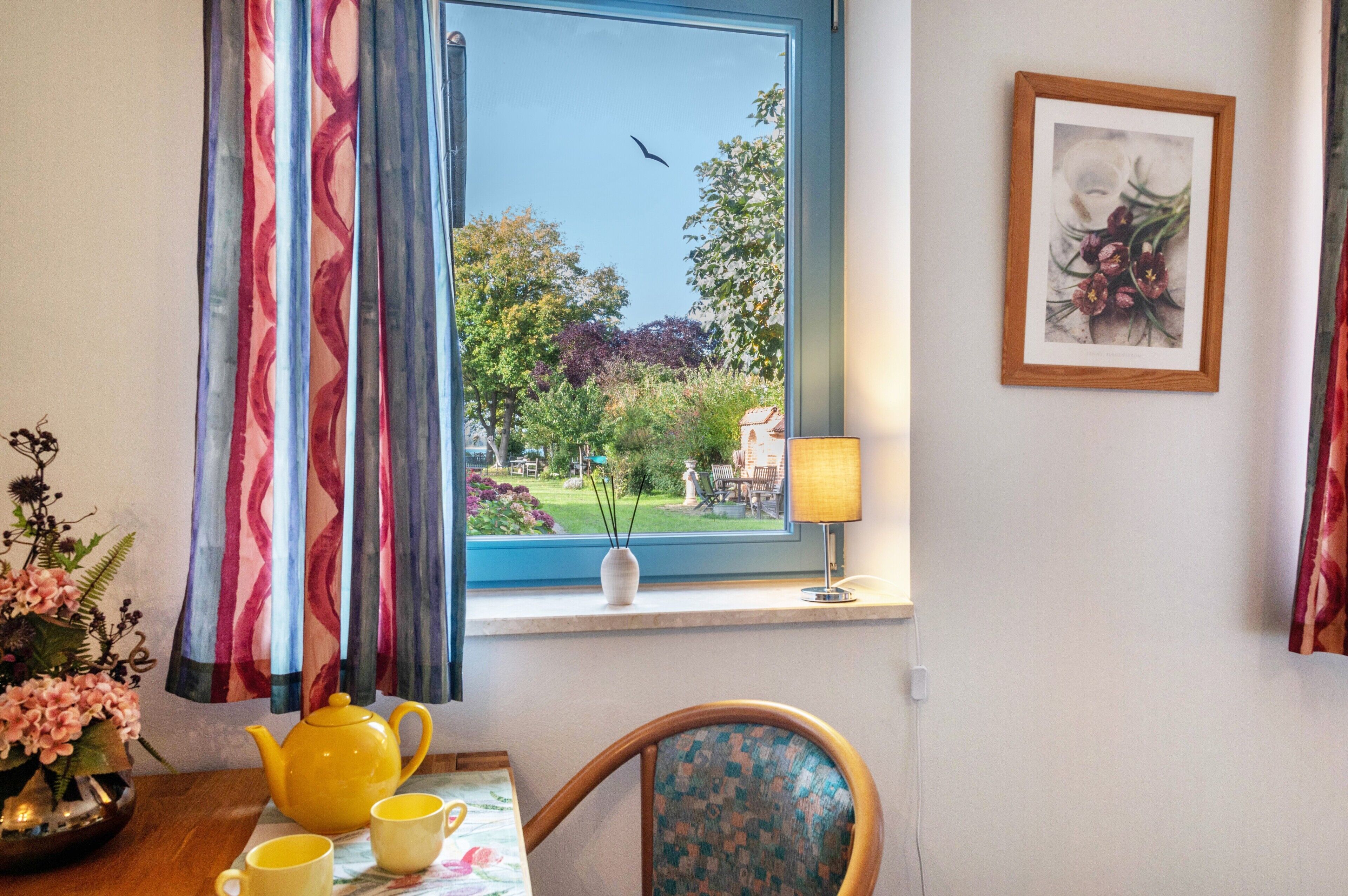Une pièce avec une fenêtre, une table sur laquelle se trouvent une théière jaune et des tasses, une chaise, une lampe et un tableau encadré accroché au mur.