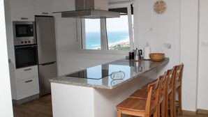 A kitchen with a view of the ocean, a microwave, an oven, and a bar area with wooden chairs.
