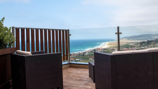 A balcony with wicker furniture, a wooden fence, and a view of the ocean and distant landscape.
