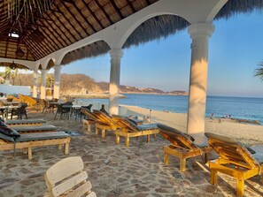 A beachside lounge area with wooden lounge chairs and tables, a thatched roof, and a view of the sea. - HOTEL LA ISLA HUATULCO H (Santa María Huatulco)