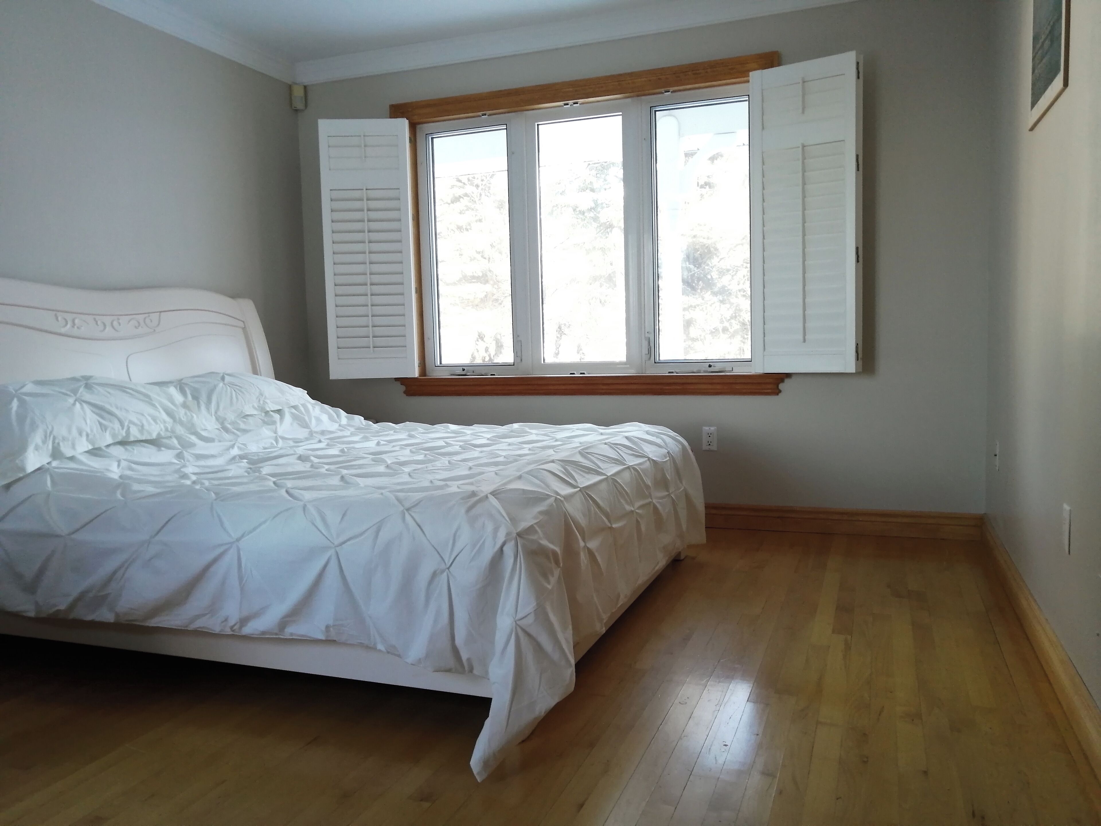 Una camera da letto con un letto, pavimento in legno, una finestra con persiane e una testiera bianca.