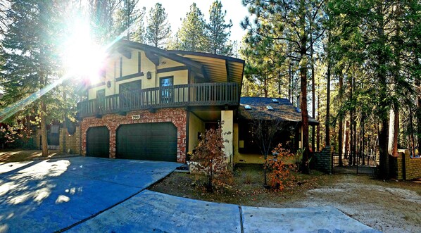Situated in a wooded area, this two-story home features a balcony and a garage, surrounded by a driveway and a small front yard with trees and shrubs.