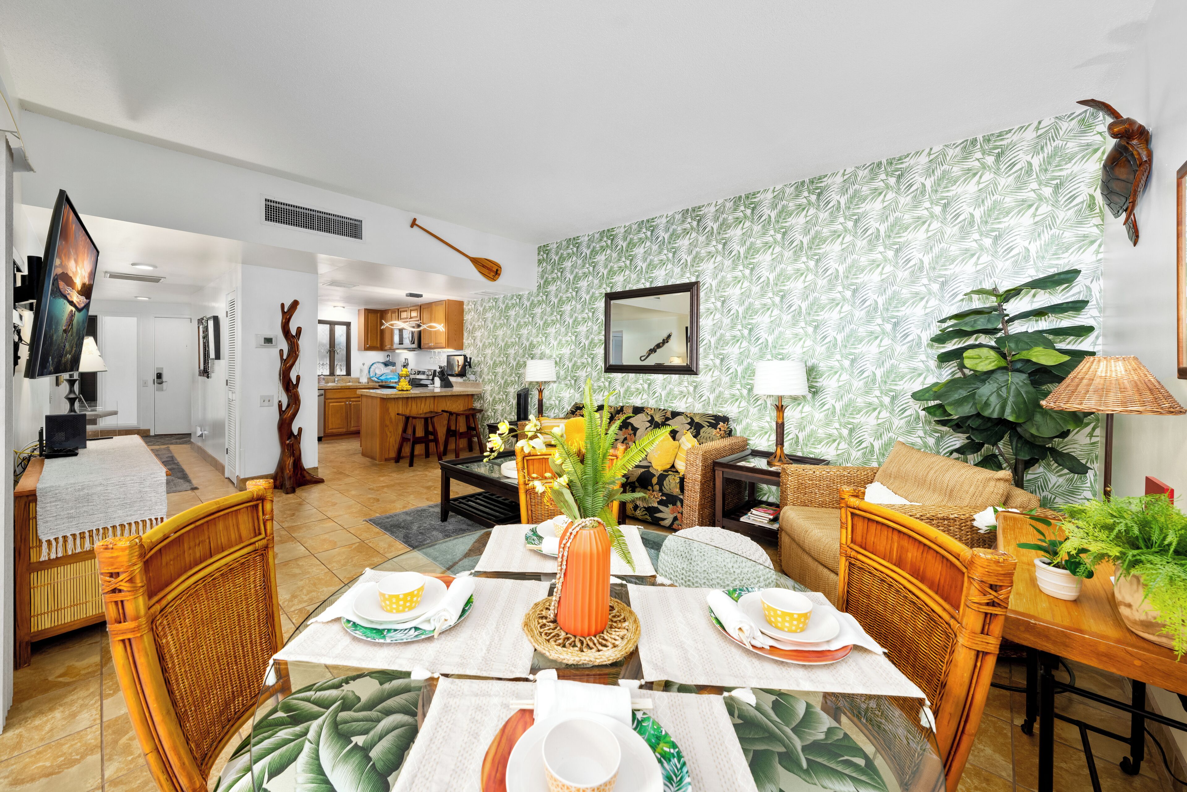 Un coin repas avec une table dressée pour quatre personnes, une cuisine en arrière-plan et un salon avec un canapé et une grande plante.