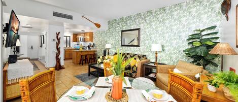 Un coin repas avec une table dressée pour quatre personnes, une cuisine en arrière-plan et un salon avec un canapé et une grande plante.