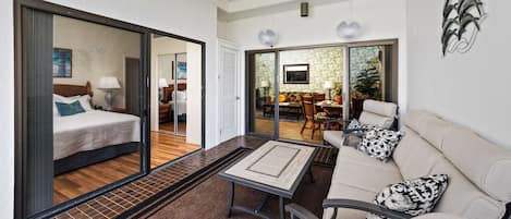 A modern hotel room with a bed, a sofa, a coffee table, and a dining area visible through a glass door.