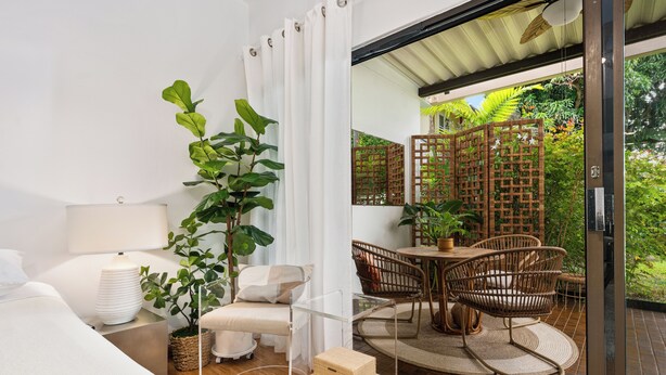 Ein modernes Schlafzimmer mit Glastür zum Balkon, einem Holztisch, Korbstühlen und einem Topfpflanzen.