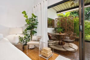 A modern bedroom with a glass door leading to a balcony, a wooden dining table, wicker chairs, and a potted plant. - Castle at Royal Sea Cliff  (Kailua-Kona)