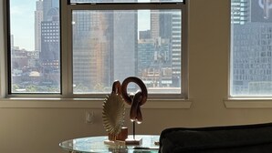 A room with a large window offering a cityscape view, a round glass table with a decorative metal sculpture, and a dark-colored sofa. - Charming 1-bedroom apartment with views (Dallas)