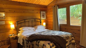A wooden cabin room with a bed, two bedside lamps, a nightstand, and a window with curtains.