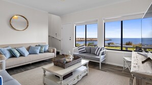 Een ruime woonkamer met een groot raam en uitzicht op zee. Er is een bank, een salontafel en een houten bijzettafeltje.