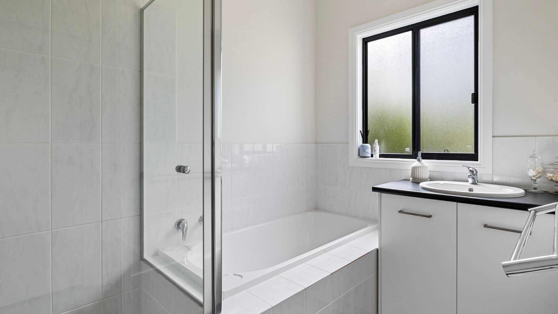 Une salle de bain moderne avec une douche à l’italienne en verre, une baignoire blanche, un lavabo avec un plan de toilette noir et une fenêtre au-dessus du miroir.