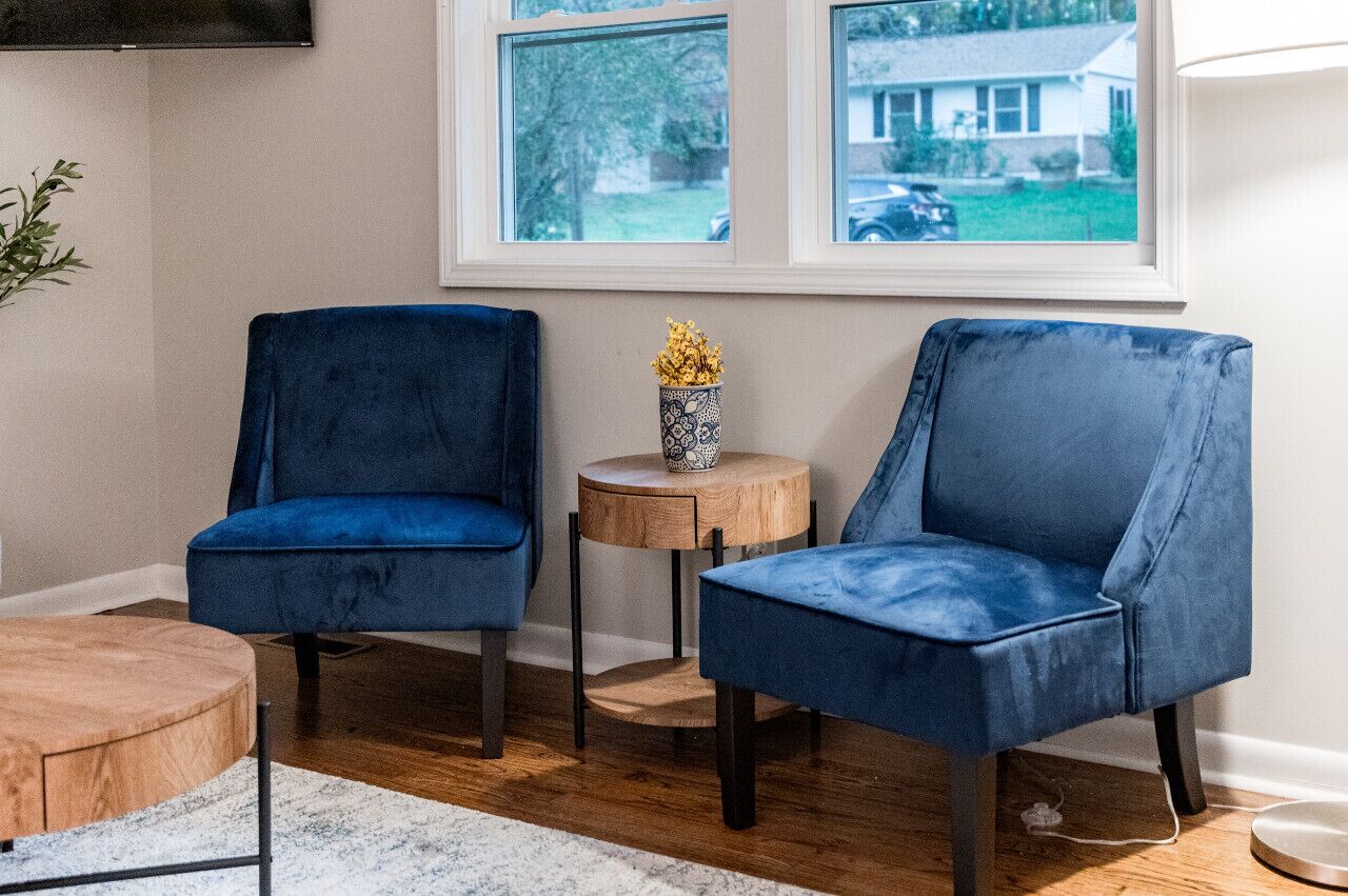 Una habitación con dos sillas azules, una mesita redonda de madera con un florero, y vista a una casa a través de una ventana.