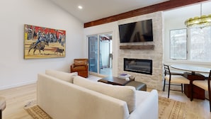 A living room with a sofa, a brown leather chair, a fireplace, a TV mounted on the wall, and a dining area with a table and chairs. - JHRL - Hollyhock 2023 (Wilson)