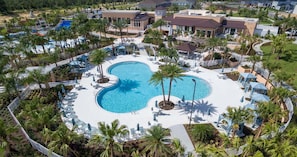 An aerial view of a resort with a central swimming pool, surrounded by palm trees and lounge areas.