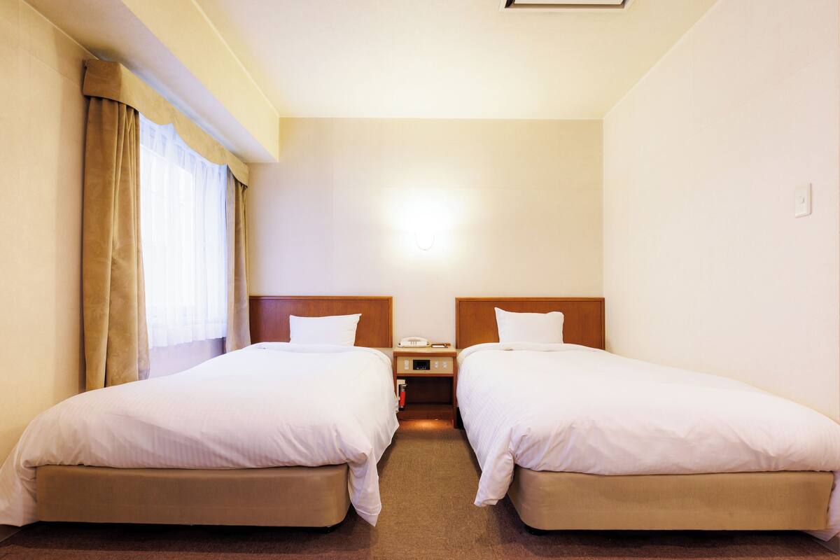 A hotel room with two single beds, a nightstand with a phone and a lamp, and a window with curtains.