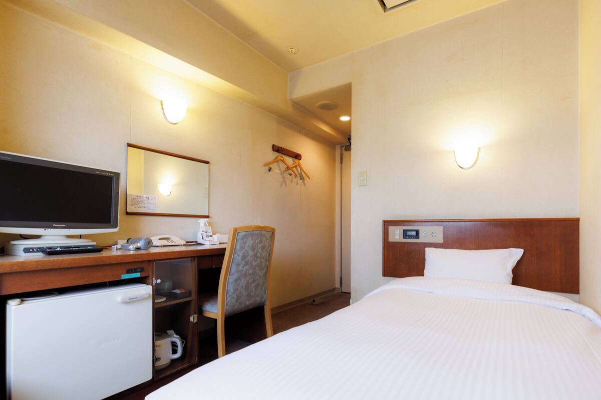 A hotel room with a bed, a desk with a computer, a chair, a small refrigerator, and a wall-mounted TV.
