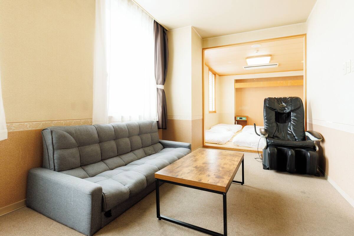A hotel room with a grey sofa, a wooden table, a bed with white linens, a black massage chair, and a small bedside table with a lamp.