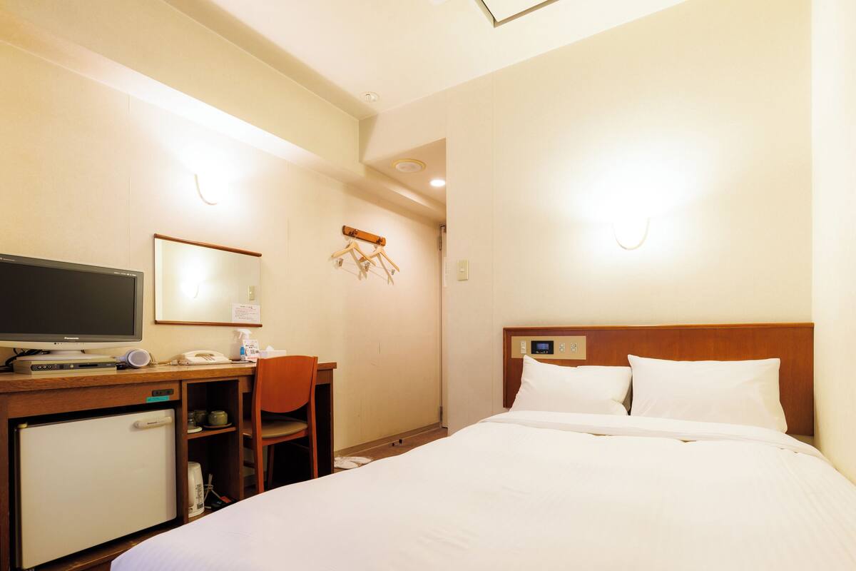 A hotel room with a bed, a desk with a computer, a chair, a small refrigerator, and a mirror.