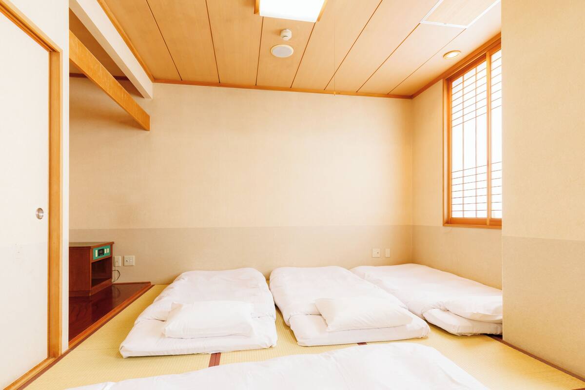 A room with two beds, a small wooden cabinet, and a window with blinds.