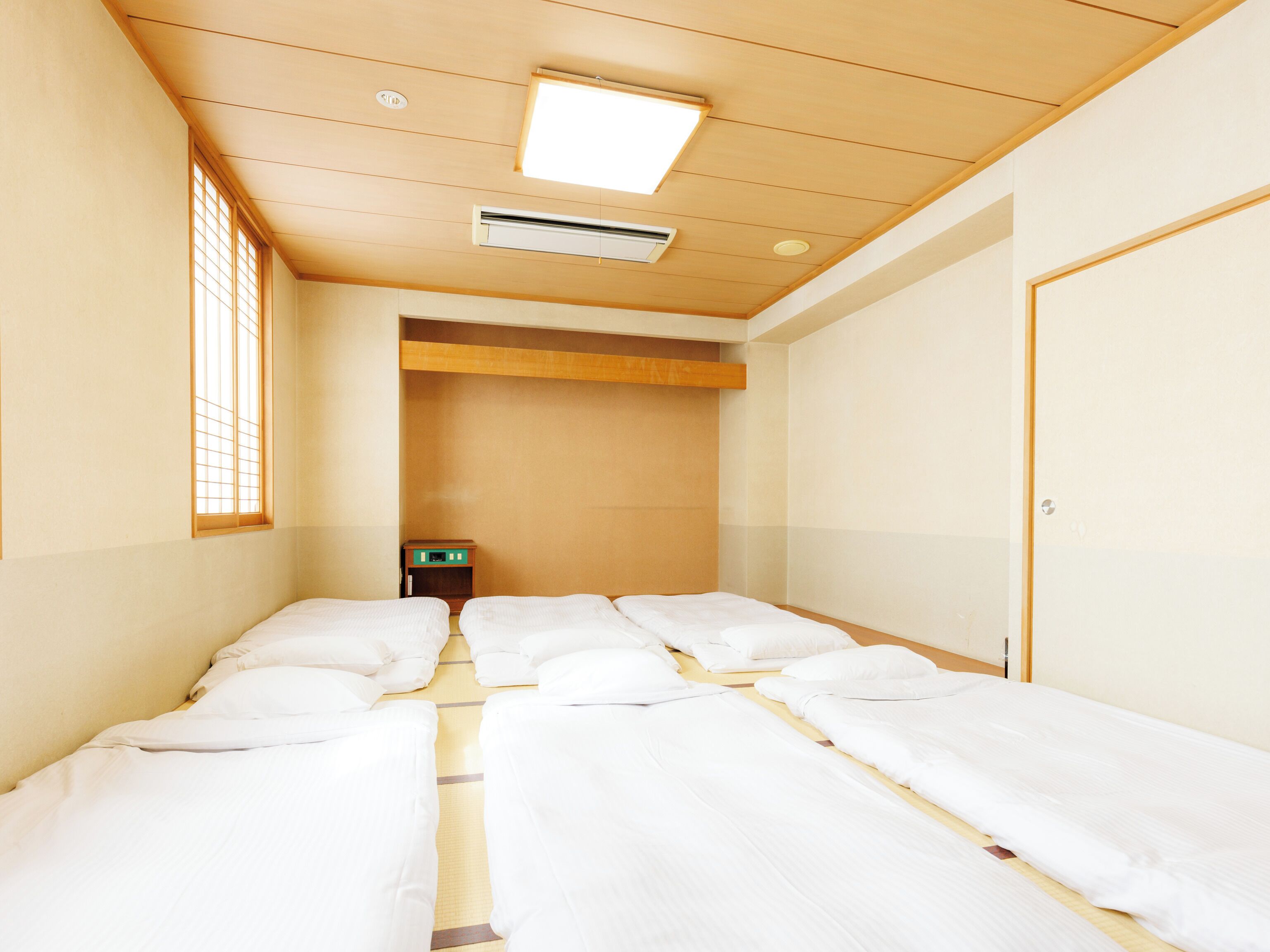 A hotel room with four single beds aligned in a row, a small bedside table, a wall-mounted air conditioner, and a window with a grid pattern.