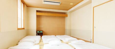 A hotel room with four single beds aligned in a row, a small bedside table, a wall-mounted air conditioner, and a window with a grid pattern.