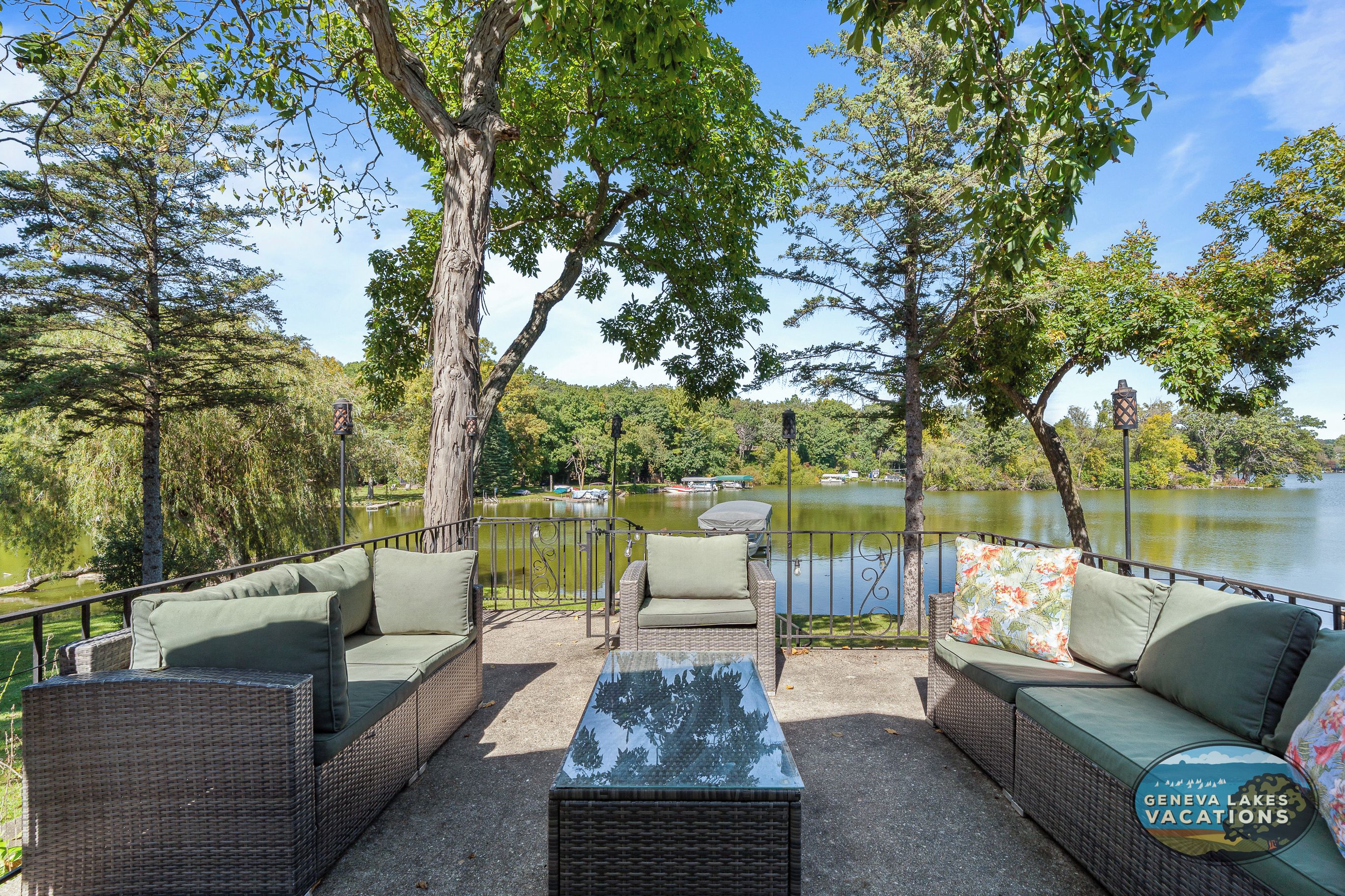 An outdoor seating area with wicker furniture and a glass-top table, overlooking a lake.