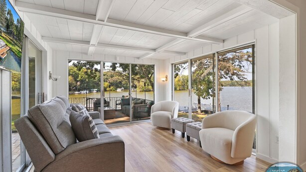 A spacious balcony with a sofa, armchairs, and a small table. It overlooks a lake with boats and trees.