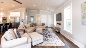 A spacious living area with a large sectional sofa, a dining area with a kitchen in the background, and a large window providing natural light. - OFF THE GRID-Custom Showplace w/ Private Pool;Multiple Decks;3 Minute Walk to Beach (Edisto Beach)