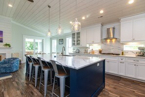 A modern kitchen with white cabinets, a large island with bar stools, pendant lighting, and a view to the outdoors. - OFF THE GRID-Custom Showplace w/ Private Pool;Multiple Decks;3 Minute Walk to Beach (Edisto Beach)