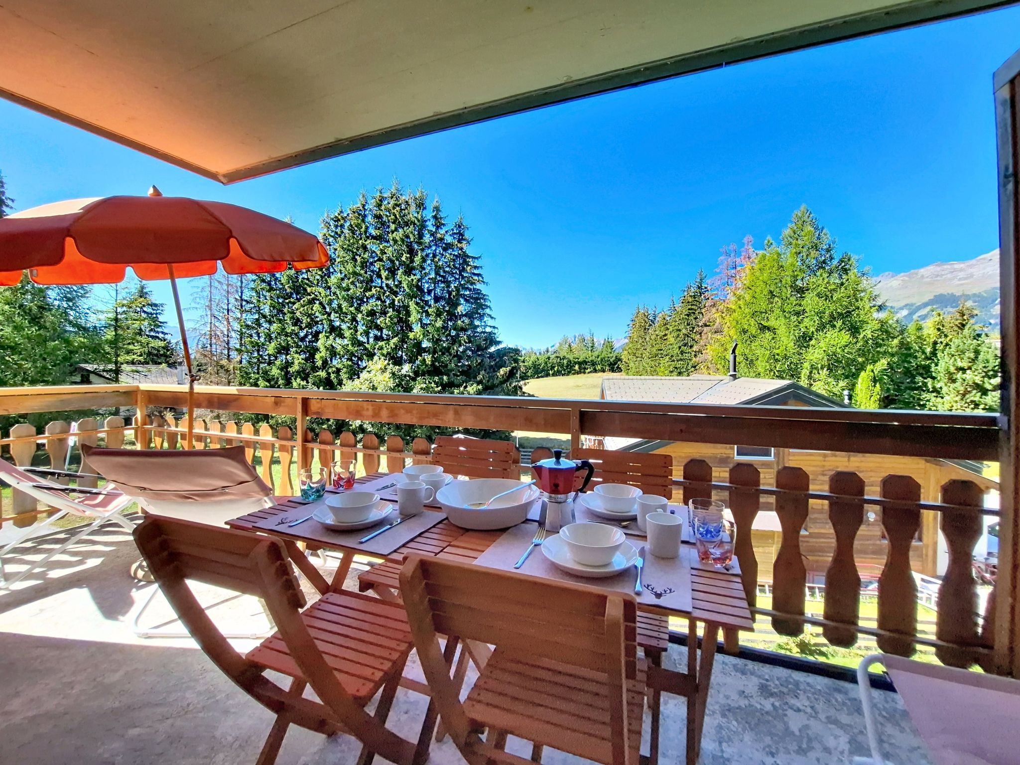 Eine Holzterrasse mit Esstisch, mit Blick auf eine bewaldete Landschaft und Berge in der Ferne.
