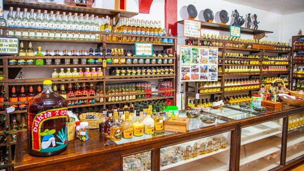 A liquor store with shelves stocked with various bottles and a display counter with a large bottle of Delicia Zapata. - Hotel del profe (Santa María Huatulco)