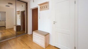A modern apartment with a wooden floor, a sliding glass door leading to a kitchen, a white door with a small wooden sign, and a light-colored bench.