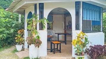 A covered outdoor seating area with a table and chairs, surrounded by potted plants.