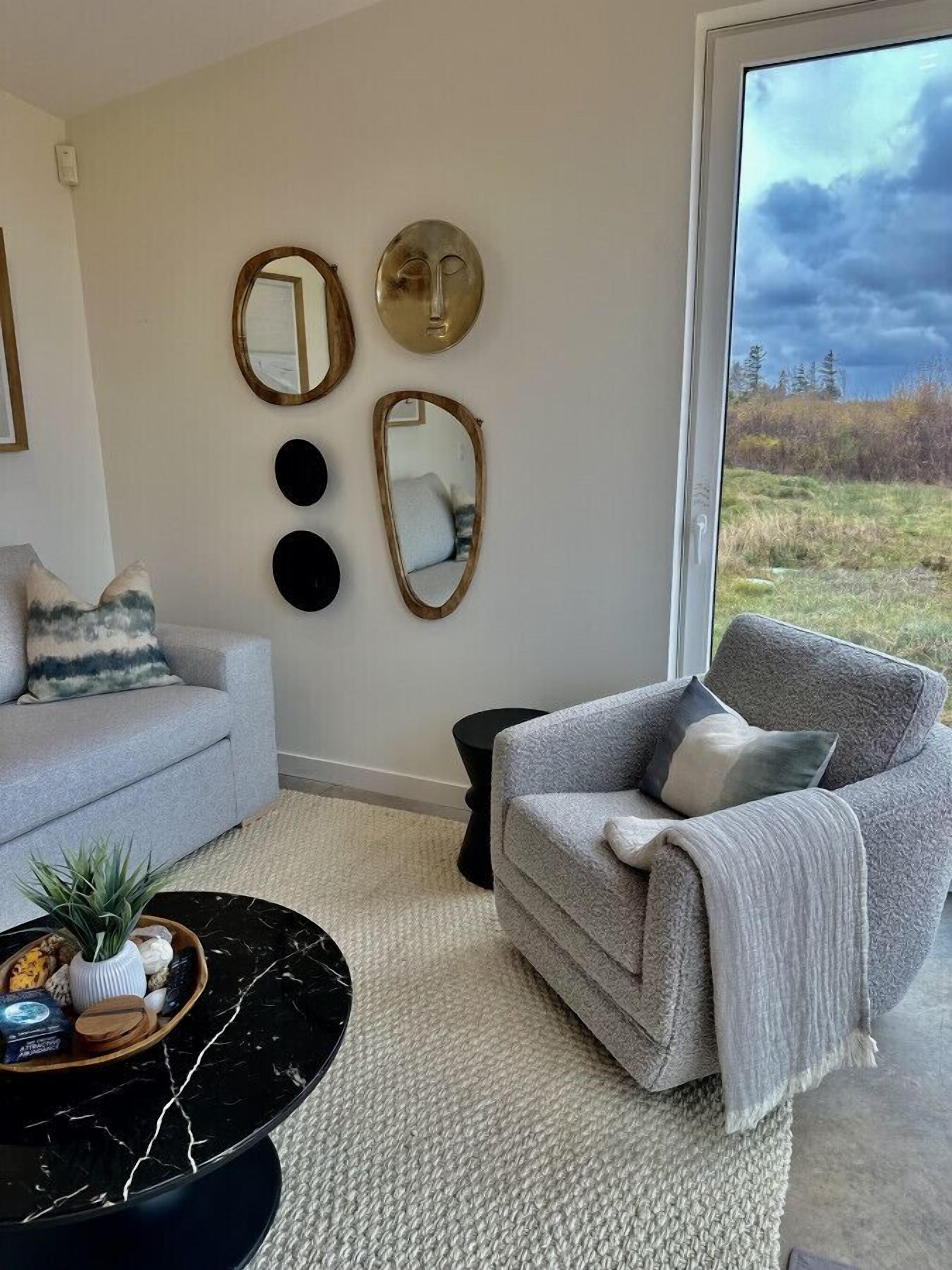 Uma sala de estar com um sofá cinzento, uma mesa de centro redonda preta e vista para uma paisagem natural através de uma janela grande.