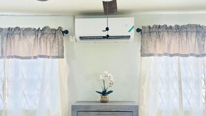 Ein Zimmer mit Deckenventilator, Klimaanlage, einer Holzkommode mit Schubladen und durchscheinenden Vorhängen.