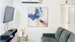Ein Wohnzimmer mit grauem Sofa, einem weißen Fernseher, einem Deckenventilator, einer Klimaanlage und einem farbenfrohen, abstrakten Gemälde.