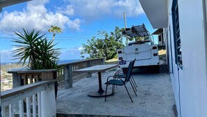 A balcony with a table and chairs, overlooking a scenic view with a truck parked nearby. - Bamboo – New Apt. - Downtown/Sea View - Perfect Location (Charlotte Amalie West)