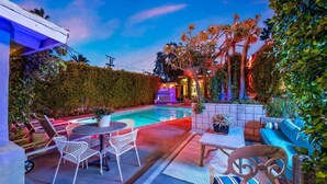 An outdoor pool area with seating and a small table, surrounded by lush greenery and palm trees. - El Mirador; Unit E (Palm Springs)