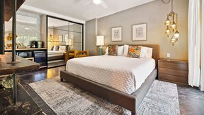 A modern bedroom with a large bed, a ceiling fan, a chandelier, and a view of a kitchen area. - El Mirador; Unit E (Palm Springs)