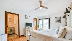 A bedroom with a wooden floor, a bed with white bedding, a ceiling fan, a TV, and a window with curtains.