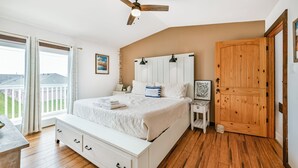 A bedroom with a large bed, wooden floor, ceiling fan, and a view of the outdoors.