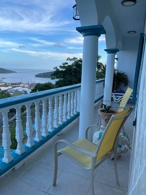 Un balcon doté de balustrades blanches et de chaises jaunes, offrant une vue sur une zone côtière où l’on aperçoit des grues de chantier et de la végétation.
